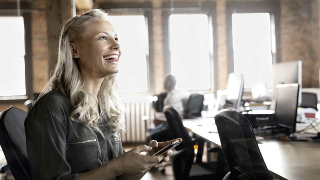 Businesswoman with cell phone laughing in office 1240 x 698