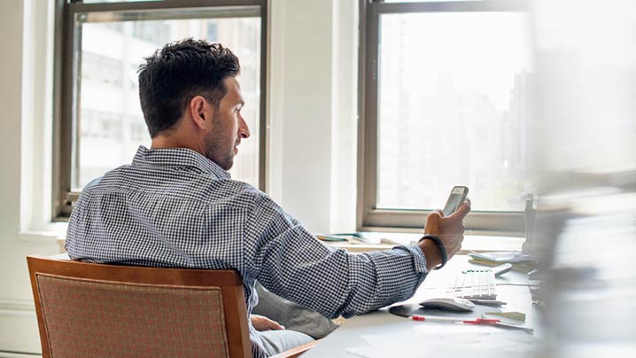 A Man At A Desk Checking His Phone 718 x 404